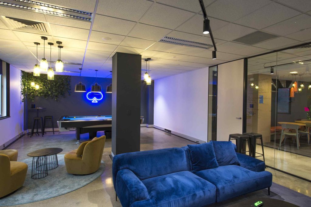 modern office design with glass wall dividing rooms, front room has velvet blue couch, rug, two fawn coloured chairs and coffee table, pool table and neon company log on wall. Second room has work area