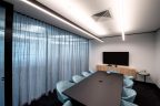 Meeting room with soft blue velvet mid-chairs, dark long table and tv at end on light wood bench. Transparent curtains on one wall with glass and light behind.