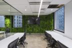 Back wall covered in plants with large screen on wall, marble style flooring, white long office designs, grey chairs for office staff.