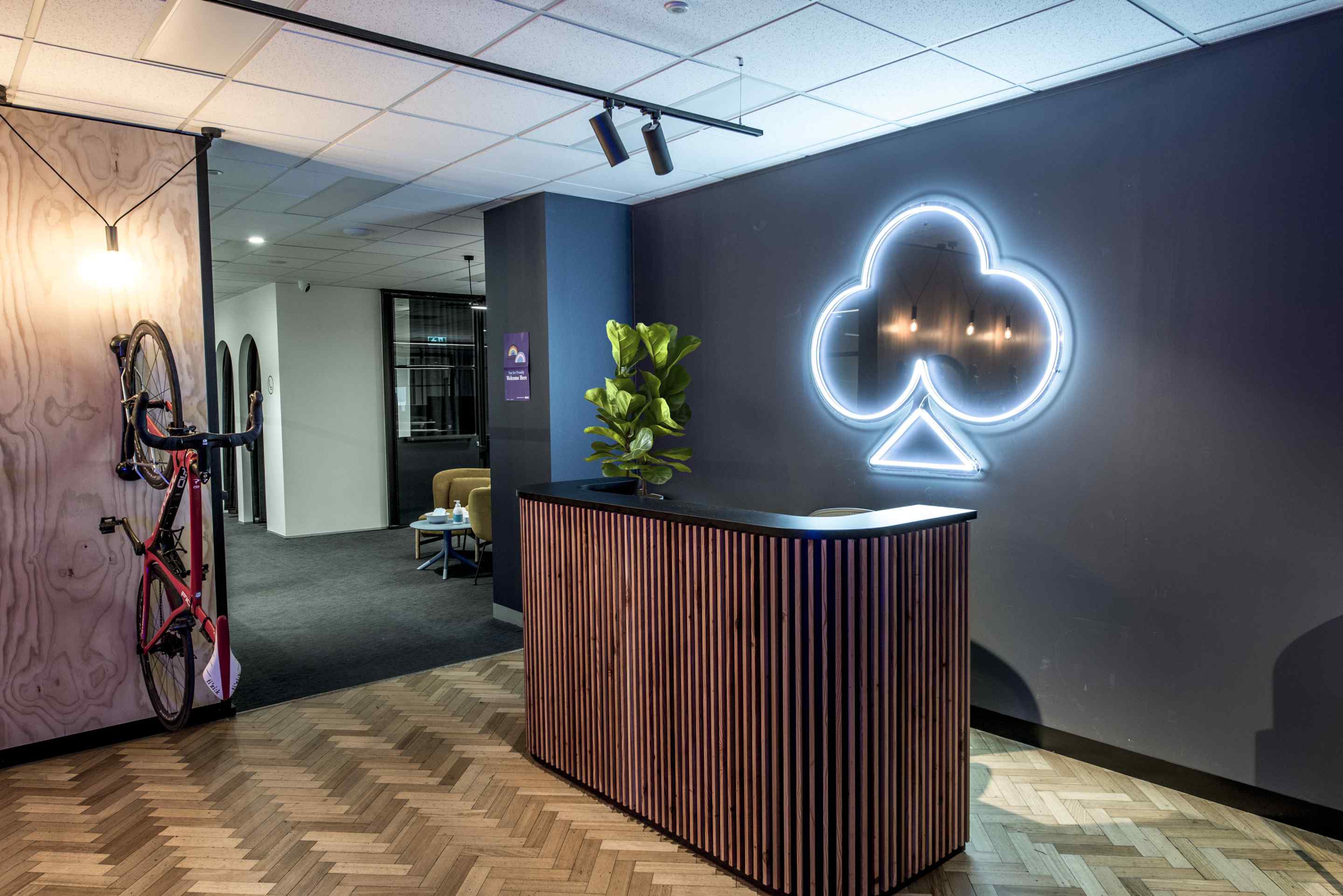 office reception design with verticle wooden slats desk with black top, illuminated logo behind on dark gray wall, red bike hung on left and entry into main office on left