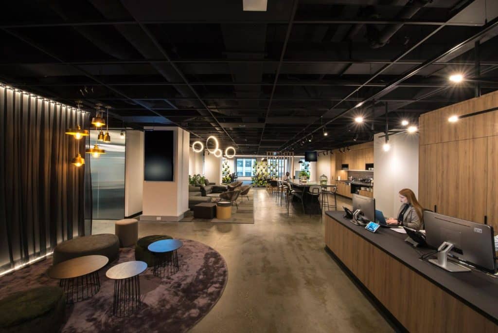 best office fitout using black exposed ceiling, long black bench top and wood accent reception area, velvet style rug in seating area, modern lighting and greenery