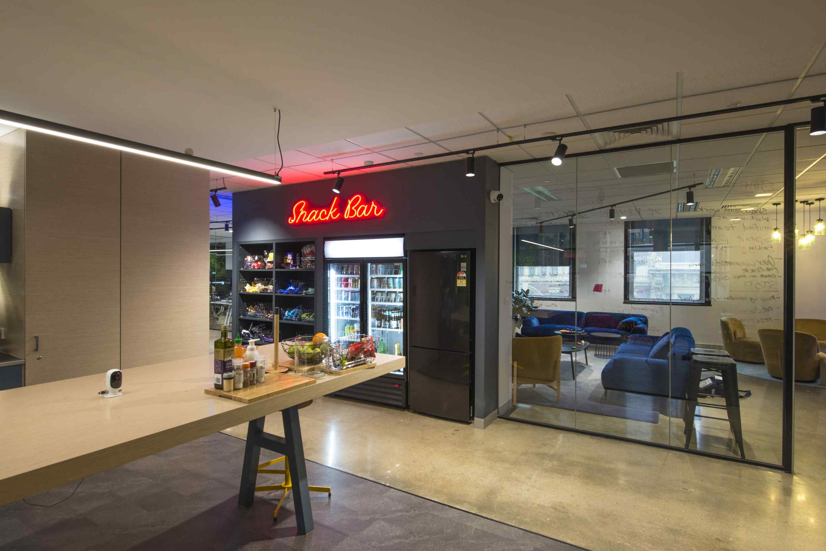 cafe style drinks fridge and open pantry with neon snack bar sign, long wooden kitchen bench, glass partitions with staff lounge in background with velvet chairs in blue and fawn colours
