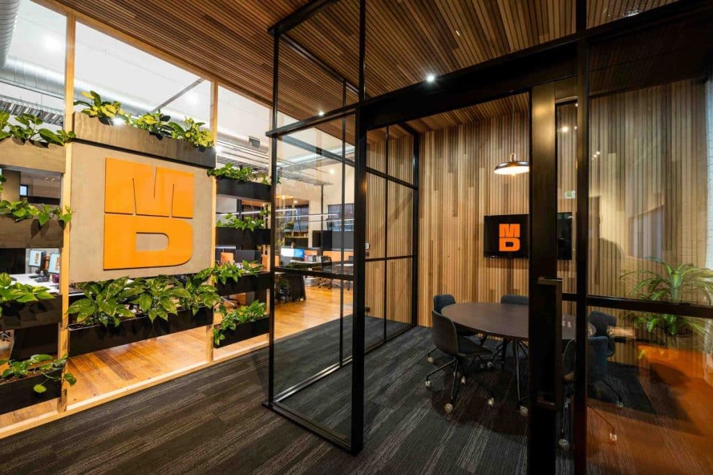 Using the company brand on two panels surrounded by plants, charcoal panel carpet, meeting room with black window frames and dark desk. Wood ceiling panel and flooring