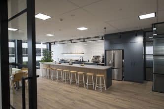 large open kitchen with long centre bench and wood accents, bench stools, white rectangle tiles above sink area, storage and fridge on right, wooden floorboards, modern and clean