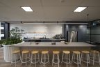 Office fitout showing beautiful, modern kitchen, long light coloured bench with bench stools, paneled planter on left of bench, kitchen in background with white shiny tiles, fridge on right & storage cupboard. Light wood-look floorboards.