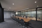 Commercial office design showing boardroom. Long wooden table to seat 14, black chairs, glass square windows with black frames, glass privacy film, grey carpet, light grey curtain at one end of privacy during meetings